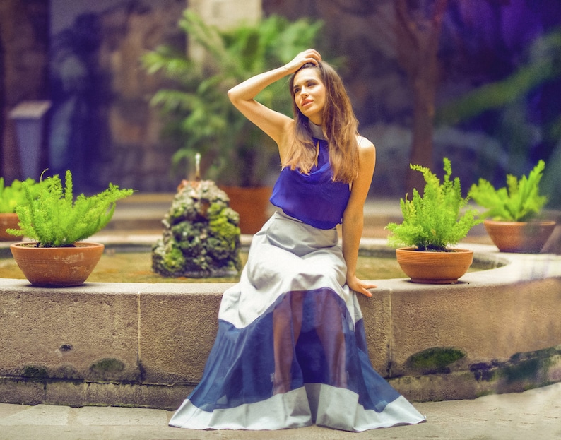May include: A woman wearing a blue top and a white and blue skirt sits on a stone ledge by a fountain. She has long brown hair and is looking to the side. The fountain is surrounded by greenery.