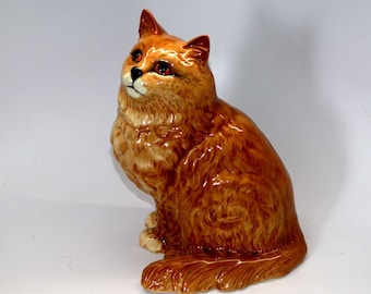 May include: A ceramic figurine of a ginger cat sitting with its head tilted up. The cat has a fluffy coat and brown eyes.