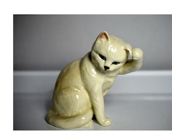 May include: A white ceramic figurine of a cat sitting with its paw raised to its head, as if scratching behind its ear. The cat has blue eyes and a light brown nose.
