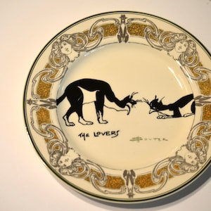 May include: A white ceramic plate with a gold and green border. The plate features a black and white illustration of two cats with the text "THE LOVERS" and the artist's signature.