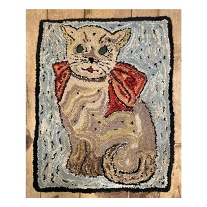 Op de afbeelding: Een handgehaakt tapijt met een beige kat met een rode strik die op een blauwe achtergrond zit.