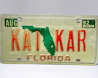 May include: A white Florida license plate with the word "KATKAR" in red letters and a green outline of the state of Florida. The plate also has the month "AUG" and the year "92" printed on it.