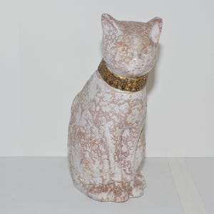 May include: White ceramic cat figurine with a gold-coloured collar. The cat is sitting with its head tilted to the side.