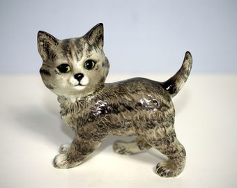 May include: A ceramic figurine of a grey tabby kitten with green eyes. The kitten is standing on all fours with its tail curled up behind it.