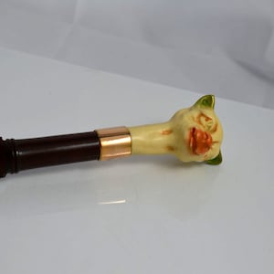 May include: A wooden walking stick with a gold band and a whimsical cat head handle. The cat head is painted in shades of cream, orange, and green.