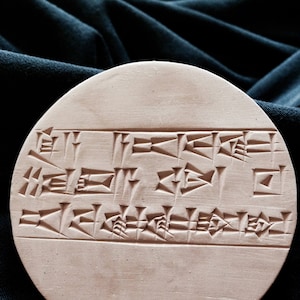 May include: A round, light brown clay tablet with cuneiform script carved into the surface.