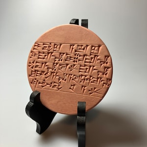 May include: A round, terracotta clay tablet with cuneiform script, displayed on a black stand.