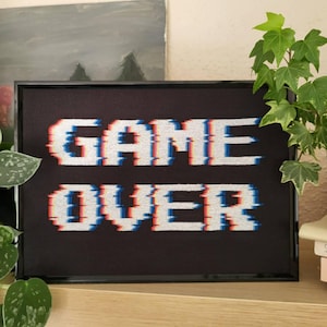 Puede incluir: Una obra de arte en punto de cruz enmarcada con las palabras "GAME OVER" en blanco, con efectos de glitch rojos y azules, sobre un fondo negro. La obra está en un marco negro y se encuentra sobre una estantería de madera, con plantas en el fondo.