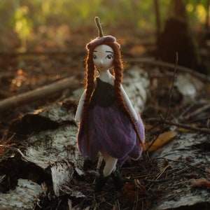 May include: A handmade doll with purple fabric clothing and brown hair, standing in a forest setting.