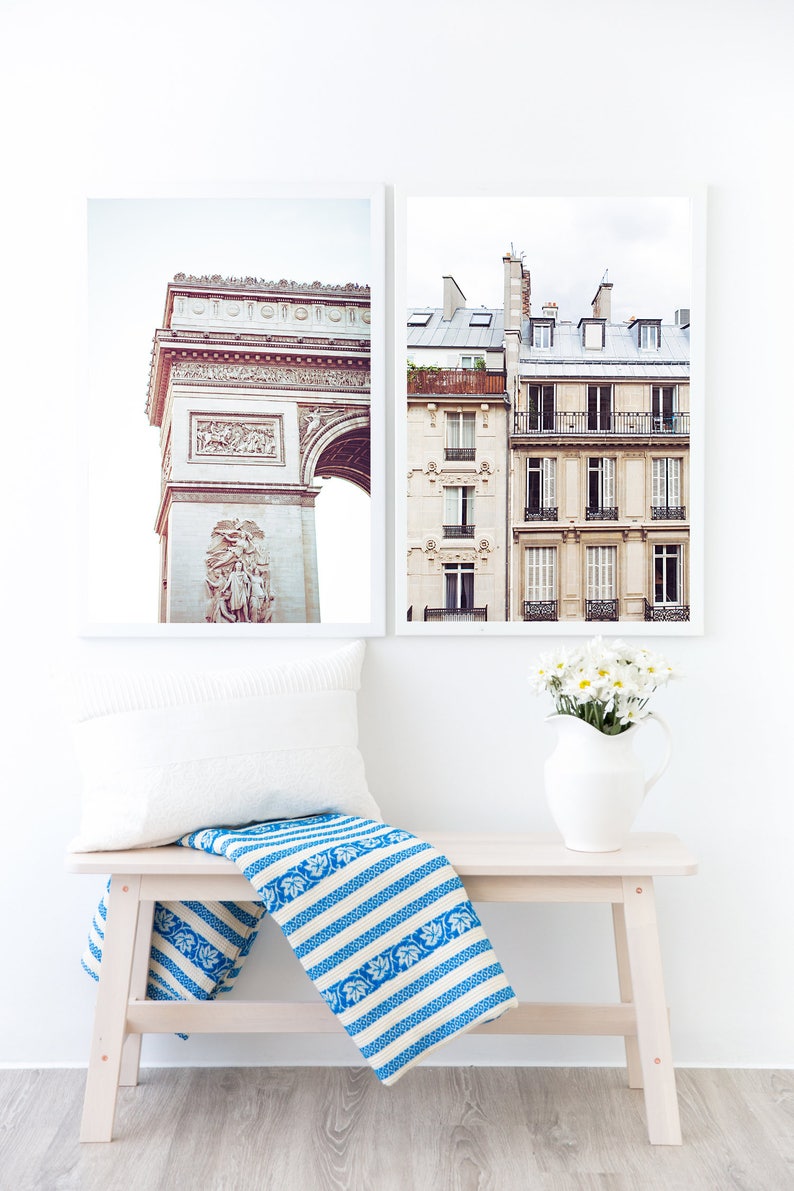 May include: Two framed photographs of Parisian architecture. The first photo shows the Arc de Triomphe, and the second photo shows a building with balconies and windows. A white bench with a blue and white striped throw blanket sits in front of the photos.
