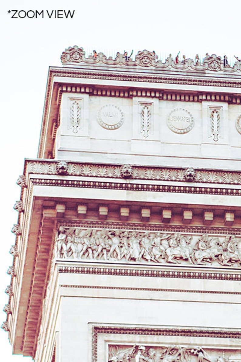 May include: A close-up view of the top of the Arc de Triomphe in Paris, France. The white stone archway is decorated with intricate carvings and sculptures. The top of the arch is adorned with a row of statues and a railing.