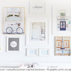 May include: A collection of framed art prints in a Parisian theme, including a bicycle, a shop front, and the word "Amour". The prints are in white frames and arranged on a white wall, with gold sconces.
