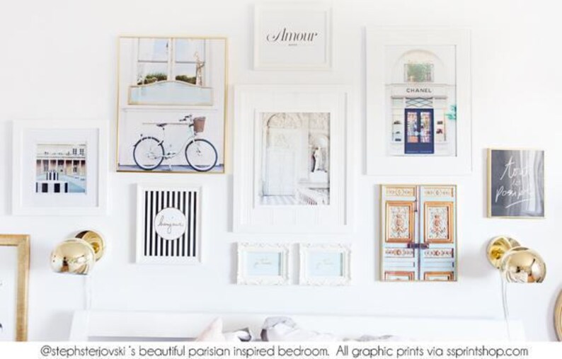 May include: A collection of framed art prints in a Parisian theme, including a bicycle, a shop front, and the word "Amour". The prints are in white frames and arranged on a white wall, with gold sconces.
