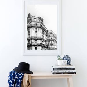 May include: A black and white photograph of Parisian buildings framed in a white frame, hanging on a white wall above a wooden bench. The bench has a stack of books and magazines on it, including a book titled "Dior Glamour".