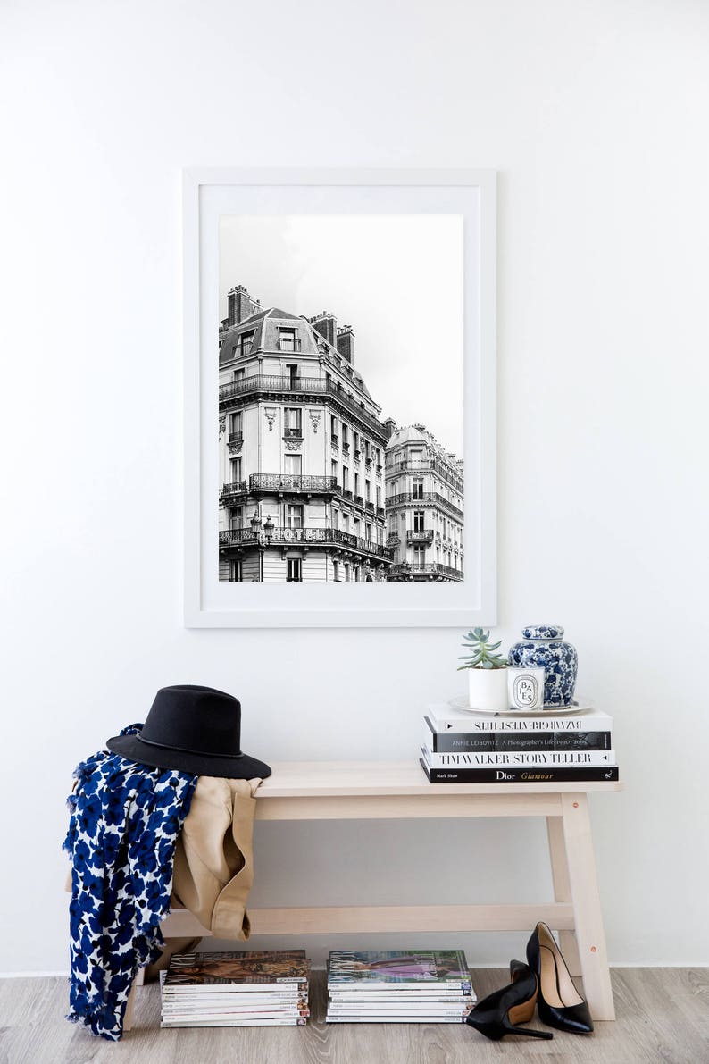 May include: A black and white photograph of Parisian buildings framed in a white frame, hanging on a white wall above a wooden bench. The bench has a stack of books and magazines on it, including a book titled "Dior Glamour".