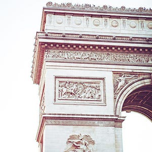 May include: A close-up of the Arc de Triomphe in Paris, France. The arch is made of white stone and is decorated with intricate carvings. The top of the arch is visible, with people standing on it.
