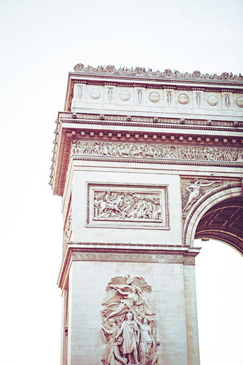 May include: A close-up of the Arc de Triomphe in Paris, France. The arch is made of white stone and is decorated with intricate carvings. The top of the arch is visible, with people standing on it.