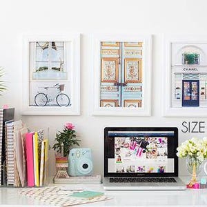 May include: Three framed prints, size 8x12 inches, depict a bicycle, ornate doors, and a shop front. A laptop, desk lamp, and stationery sit on a white desk. A small camera and flowers are also present.