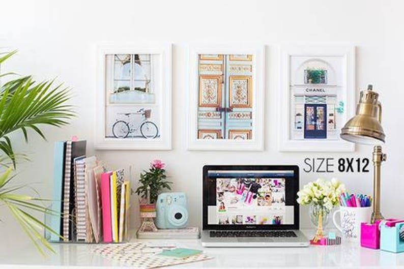 May include: Three framed prints, size 8x12 inches, depict a bicycle, ornate doors, and a shop front. A laptop, desk lamp, and stationery sit on a white desk. A small camera and flowers are also present.