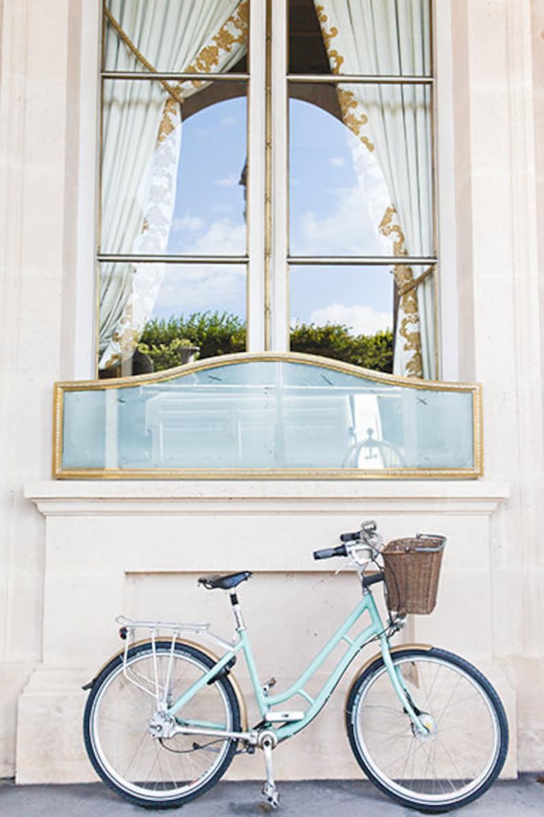 May include: A light blue bicycle with a wicker basket and a rear rack is parked in front of a window. The window has a gold-framed mirror below it and white curtains with gold accents. The scene is set against a light-colored building.