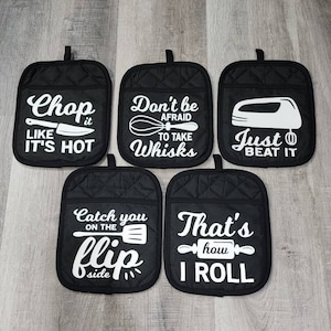 May include: Set of six black oven mitts with white text and images. The mitts feature kitchen-themed sayings such as "Chop it like it's hot", "Don't be afraid to take whisks", "Just beat it", "Catch you on the flip side", and "That's how I roll".