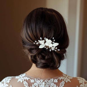Puede incluir: Una peineta floral blanca con detalles de perlas y cristales. La peineta presenta delicadas flores de tela y cristales brillantes, ideal para peinados de novia o de ocasiones especiales. El accesorio para el cabello se encuentra sobre cabello castaño oscuro.