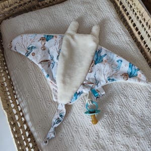 Pode incluir: Um cobertor para bebé em forma de manta-raia, com um padrão subaquático branco e azul com cavalos-marinhos e corais. O cobertor tem um centro macio e branco e uma chupeta azul presa.