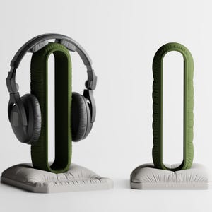 May include: Two headphone stands. One holds black headphones. The stands are olive green and sit on a white, rectangular base. The stand is U-shaped with a textured exterior. The other stand is empty.