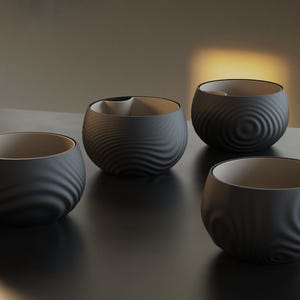 Puede incluir: Cuatro cuencos de cerámica negros con un patrón texturizado y ondulado en el exterior. El interior de cada cuenco es de color beige claro. Cada cuenco tiene una pequeña hendidura curva en el borde.