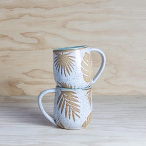 May include: Two ceramic mugs stacked. The white mugs have brown leaf patterns and a speckled texture. The inside rim of each mug is light blue. Each mug has a curved handle. The mugs are suitable for hot drinks.