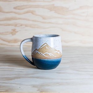 Puede incluir: Una taza de cerámica blanca con un esmalte moteado y un diseño de escena de montaña. La montaña está pintada en marrón y blanco, y la taza tiene un esmalte azul en la parte inferior.