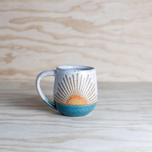Puede incluir: Una taza de cerámica blanca con un esmalte moteado presenta un diseño de amanecer pintado a mano en tonos naranja, amarillo y marrón. La taza tiene un borde y una base azul turquesa.