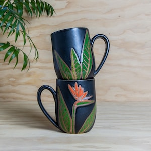 May include: Two black ceramic mugs stacked on top of each other. Each mug features a hand-painted design of a bird of paradise flower with green leaves and an orange flower. The mugs have a curved handle and are set against a light wood background.