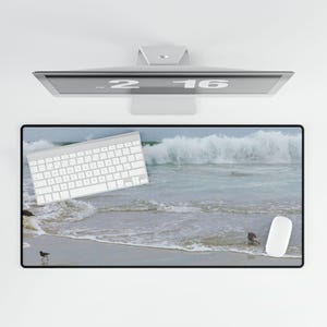 May include: A white keyboard and a white computer mouse are on a desk mat with a beach scene. The scene features a sandy beach with waves crashing on the shore. There are two birds walking on the beach.