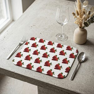 May include: A rectangular placemat featuring a repeating pattern of bright red strawberries with green leaves on a white background. The placemat is positioned on a textured surface alongside a silver fork, a silver spoon, a clear glass, and a vase.