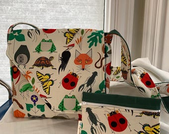 May include: A cream-colored tote bag and small pouch featuring a whimsical print of various insects, animals, and plants in red, green, yellow, and black. The tote has a green strap and flap, while the pouch has a green top and a zipper.