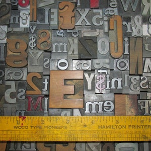May include: A collection of vintage metal and wooden letterpress printing blocks, including letters, numbers, and symbols. A yellow ruler with the text "WOOD TYPE PIONEERS" and "HAMILTON PRINTERS LINE G" is at the bottom. The letters are various sizes and fonts.