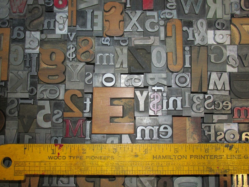 May include: A collection of vintage metal and wooden letterpress printing blocks, including letters, numbers, and symbols. A yellow ruler with the text "WOOD TYPE PIONEERS" and "HAMILTON PRINTERS LINE G" is at the bottom. The letters are various sizes and fonts.