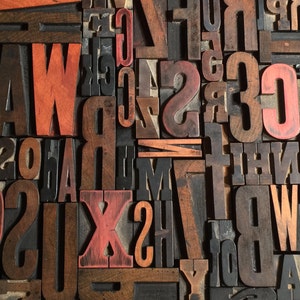 May include: A collection of vintage wooden letterpress blocks arranged in a dense, overlapping grid. The blocks feature a mix of natural dark wood tones and faded red paint, displaying various uppercase letters and numbers in different typefaces.
