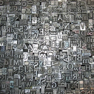 May include: A close-up of a collection of metal letterpress printing blocks. The blocks are arranged in a random pattern and include letters, numbers, and symbols.