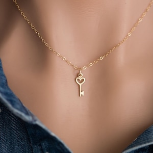 May include: A gold necklace with a delicate key pendant featuring a heart-shaped keyhole. The necklace is on a mannequin wearing a blue denim shirt.