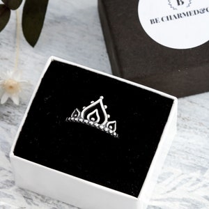 Peut inclure: Une bague en argent avec un motif de couronne dans une boîte blanche avec un intérieur noir. La boîte a un autocollant avec le texte "BE CHARMED & CO."