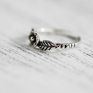 K&ouml;nnte beinhalten: Ein silberner Ring mit einem floralen Design. Der Ring hat eine kleine Blume mit einem Mittelst&uuml;ck und ein Blatt, das sich zur Seite erstreckt.