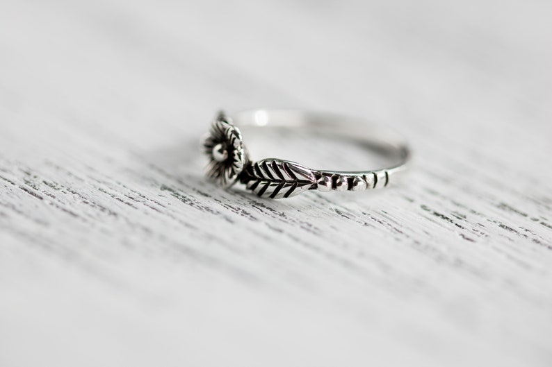 K&ouml;nnte beinhalten: Ein silberner Ring mit einem floralen Design. Der Ring hat eine kleine Blume mit einem Mittelst&uuml;ck und ein Blatt, das sich zur Seite erstreckt.