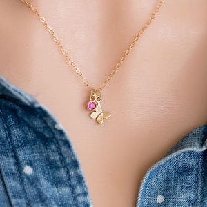 Puede incluir: Un delicado collar de cadena dorada con un pequeño colgante de mariposa adornado con una piedra preciosa rosa.