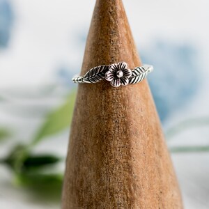 K&ouml;nnte beinhalten: Ein silberner Ring mit einem Blumen- und Blattdesign. Der Ring wird auf einem h&ouml;lzernen Ringst&auml;nder pr&auml;sentiert.