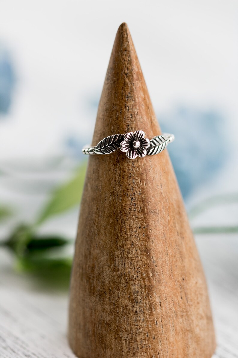 K&ouml;nnte beinhalten: Ein silberner Ring mit einem Blumen- und Blattdesign. Der Ring wird auf einem h&ouml;lzernen Ringst&auml;nder pr&auml;sentiert.
