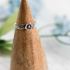 K&ouml;nnte beinhalten: Ein silberner Ring mit einem Blumen- und Blattdesign auf einem Holzringst&auml;nder.