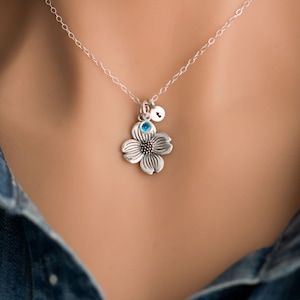 May include: A silver necklace with a delicate dogwood flower charm and a small blue gemstone. The necklace also features a small silver charm with a letter.
