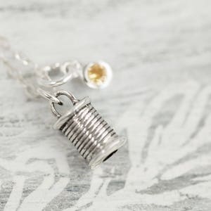 May include: Silver necklace featuring a spool of thread charm and a small yellow gemstone.